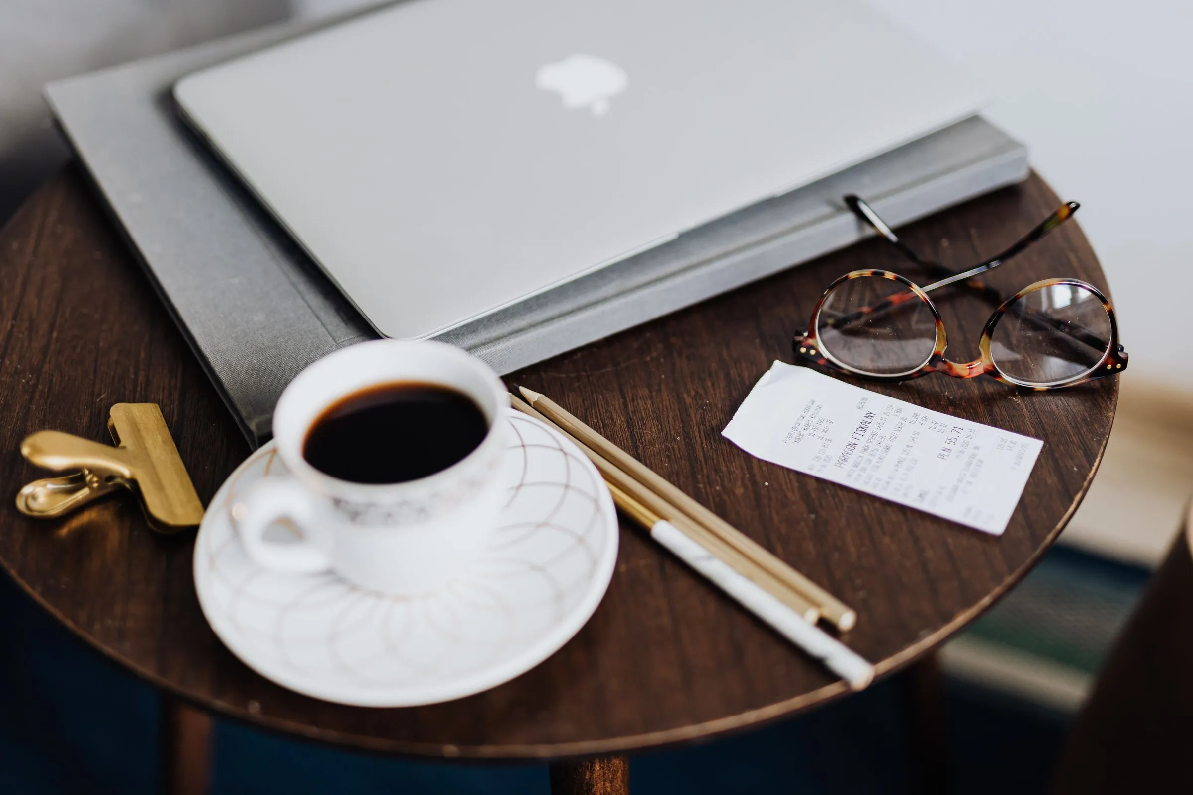 A coffee cup, a paper receipt, folded glasses and pencils on a dark wooden table beside a closed laptop