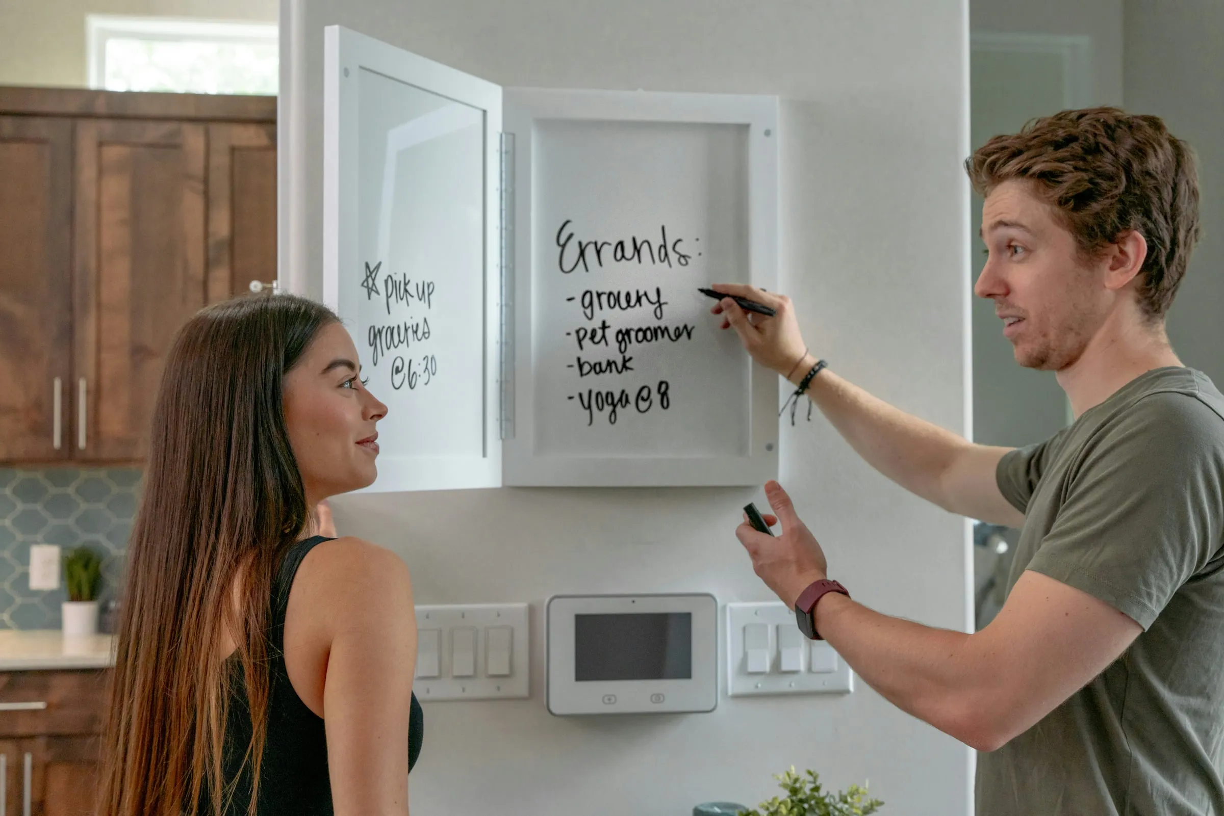 A couple at a kitchen whiteboard working out the week's errands together, one of them writing the list