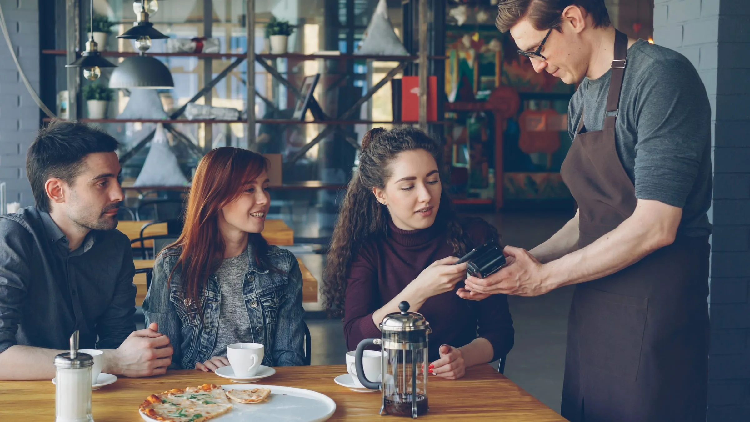 Three friends at a cafe table taking the card machine from a waiter after a meal
