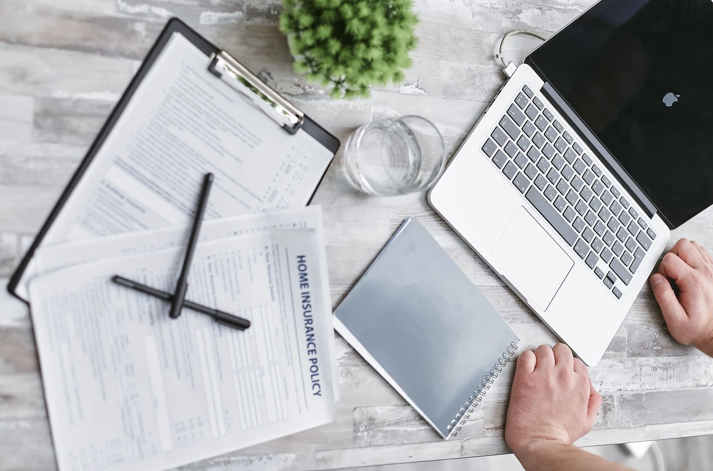 A clipboard of printed forms with a Home Insurance Policy page on top, beside a laptop, a pen and a small plant on a pale wooden table