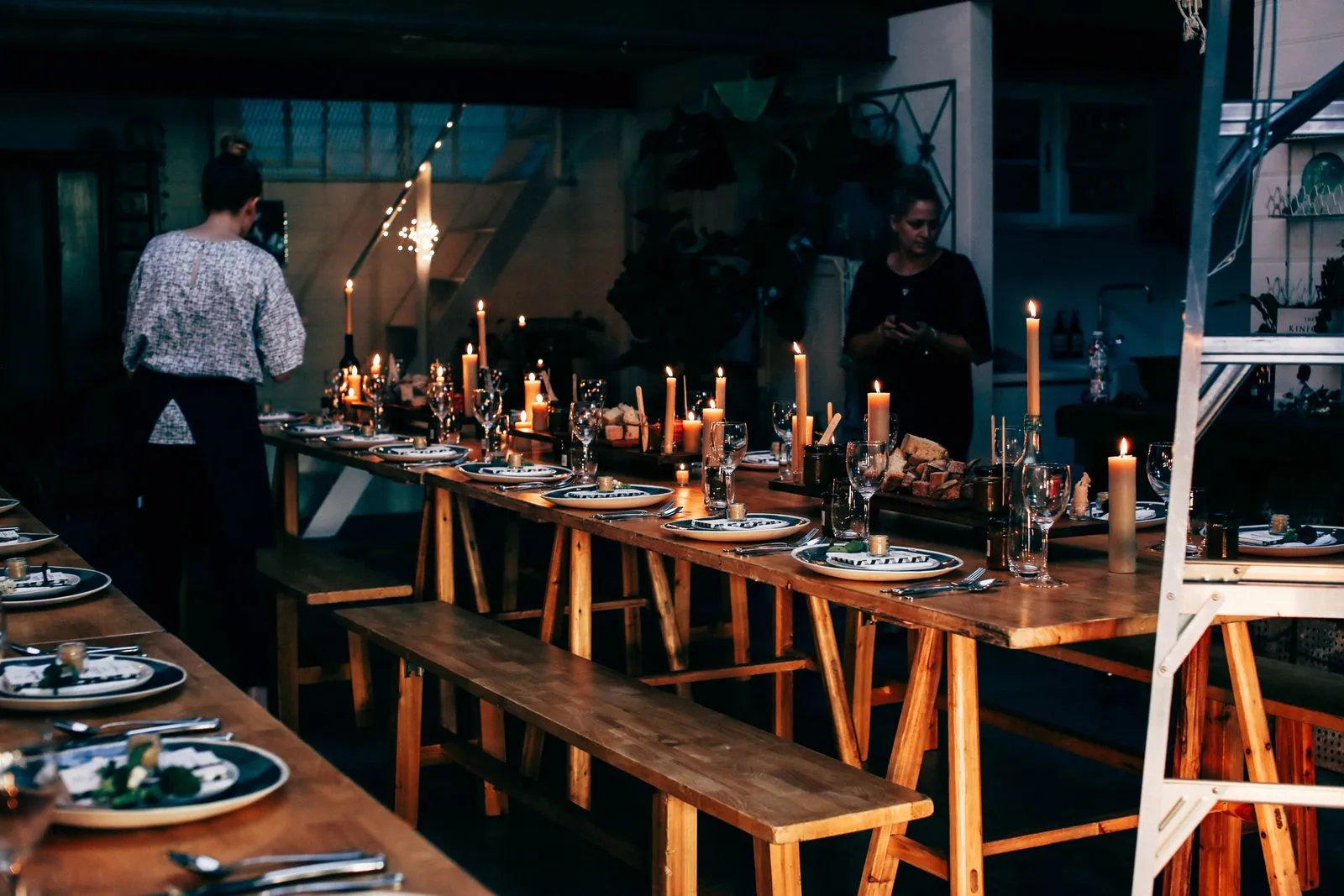 Two people finishing setting a long wooden table by candlelight before guests arrive