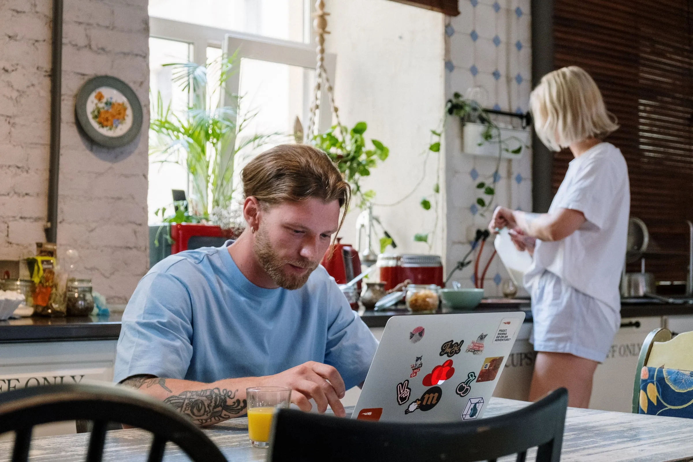 A man works at a laptop at a kitchen table while someone moves about the counter behind him, in a plant-filled, lived-in kitchen