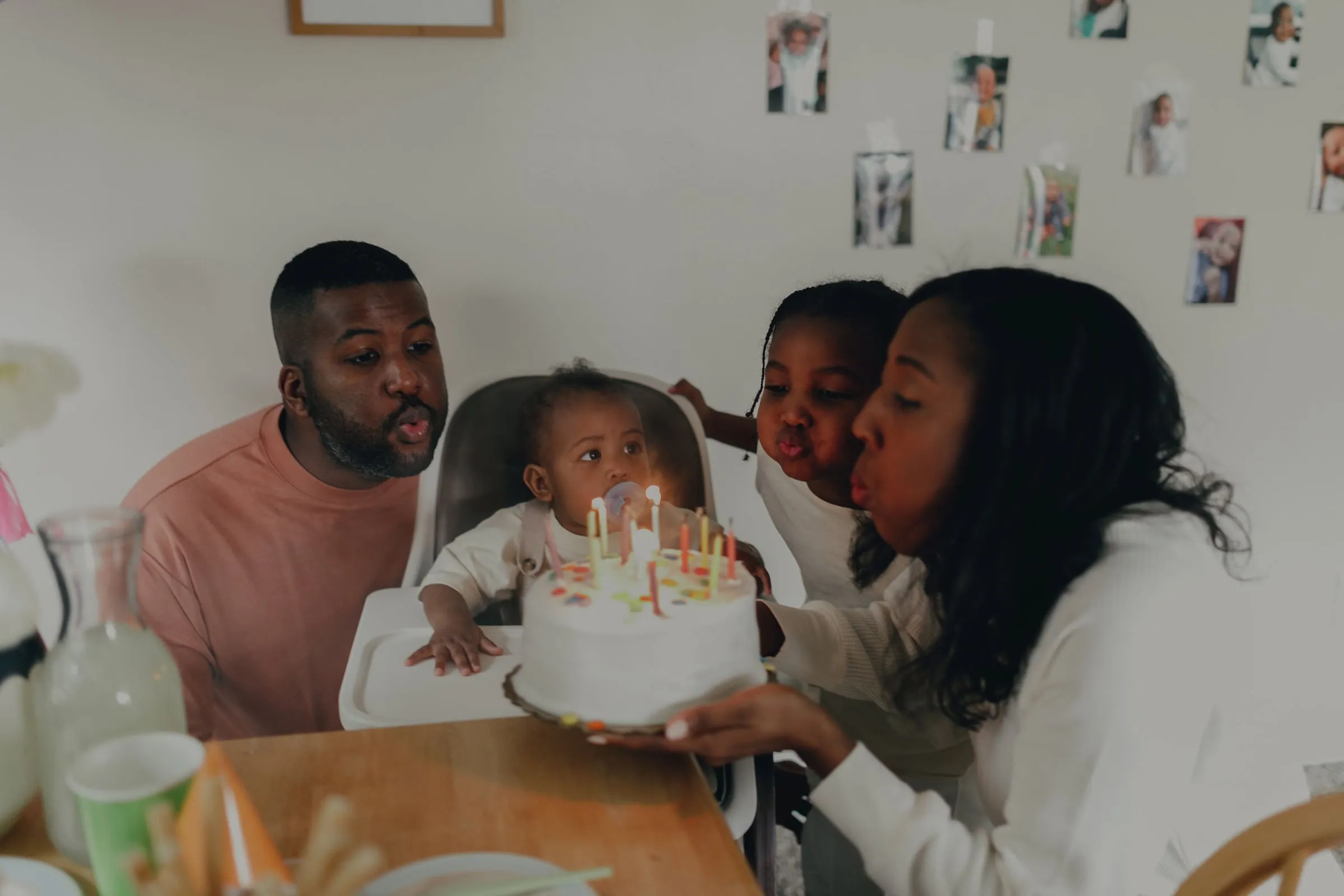 A family leaning in together to blow out the candles on a birthday cake, a baby in a high chair between them and photographs pegged along the wall behind