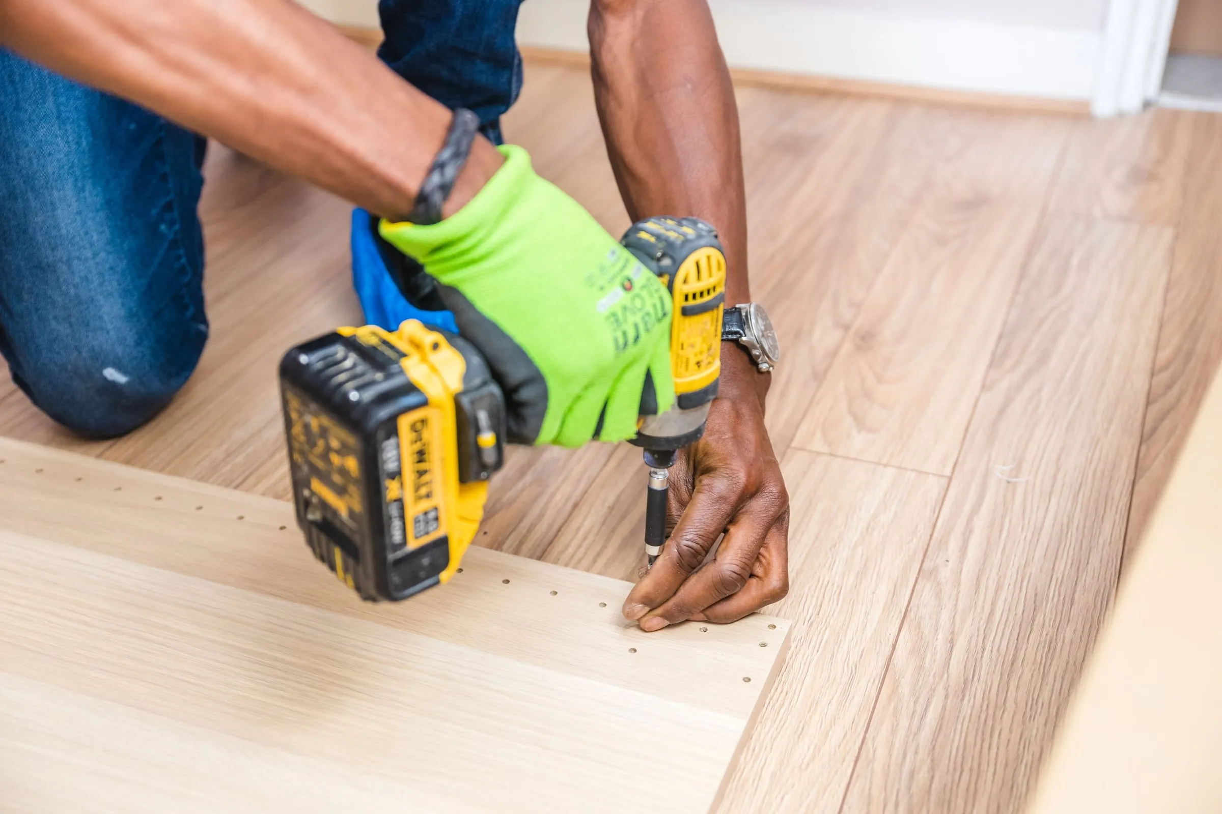 Someone kneeling on a wooden floor, driving a screw with a cordless drill